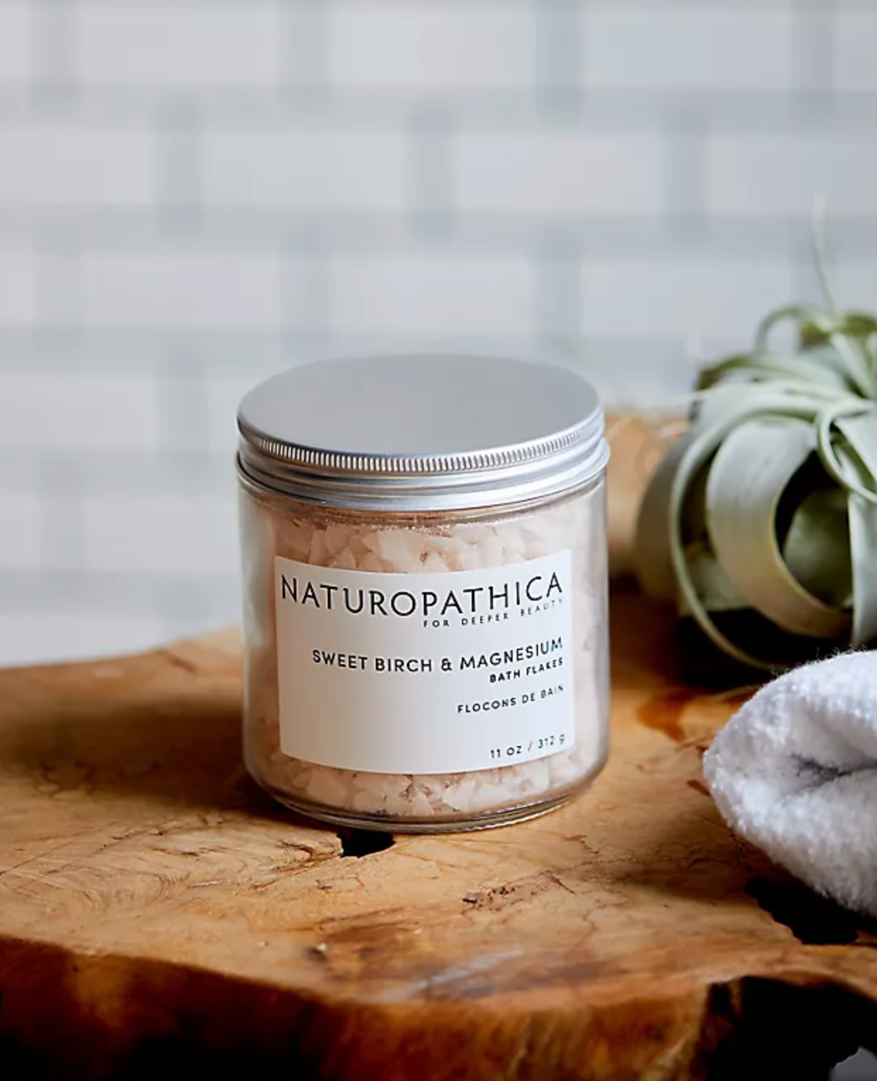 Naturopathica bath flaks jar on a wooden surface with a plant and towel in the background.